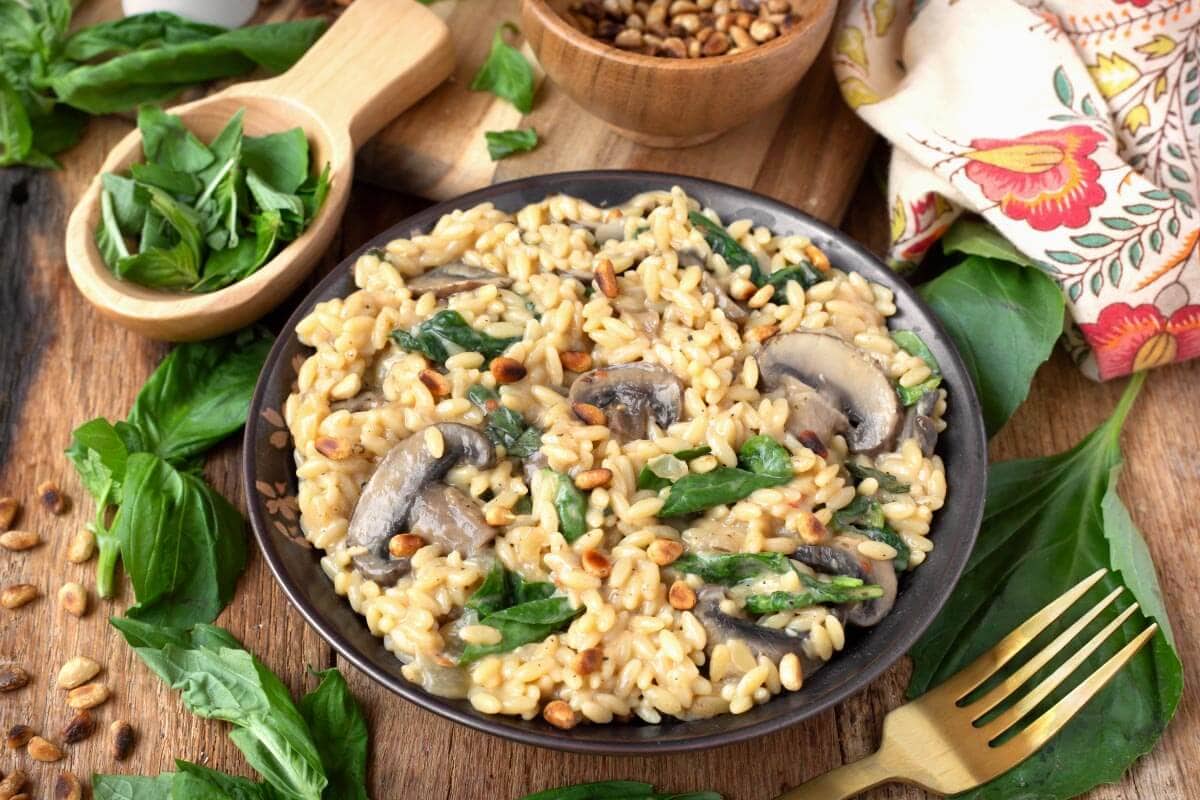 A plate of creamy orzo pasta with mushrooms, spinach, and pine nuts, garnished with fresh basil, sits on a wooden table with a fork and napkin nearby.