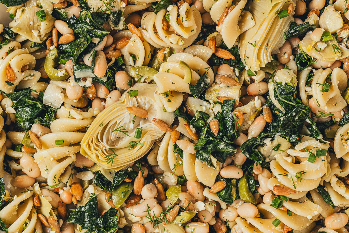 Close-up of rotini pasta mixed with artichoke hearts, white beans, cooked greens, pine nuts, and herbs.