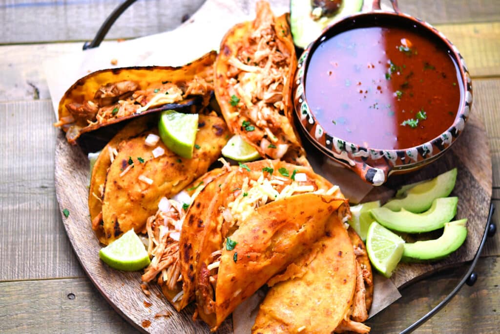 A plate of birria tacos with shredded meat, garnished with chopped onions, cilantro, lime wedges, and a bowl of red dipping sauce.