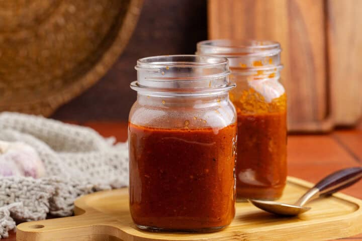 Two glass jars filled with red sauce sit on a wooden tray, with a spoon and a knitted cloth nearby.