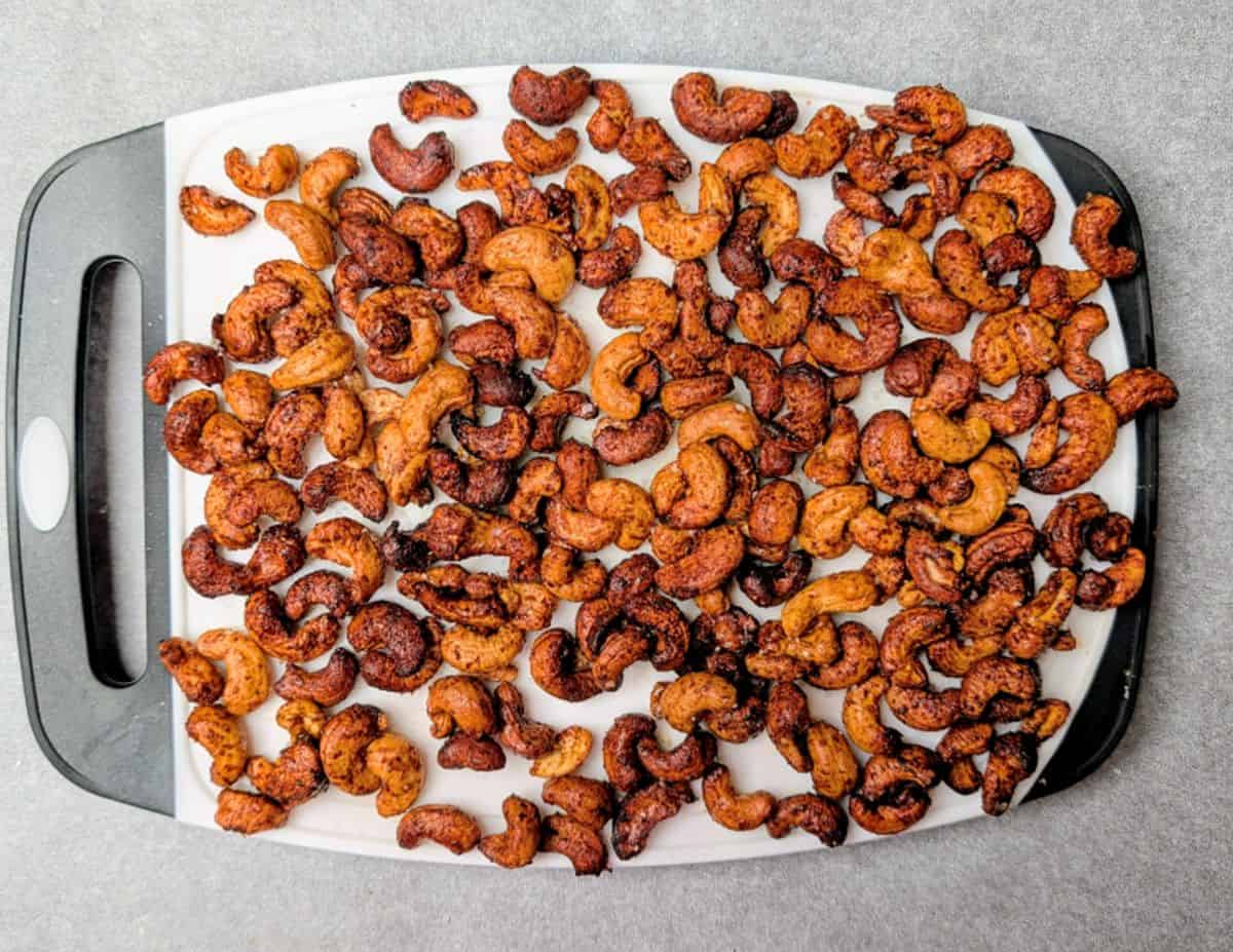 A black and white cutting board covered with a spread of roasted, seasoned cashew nuts on a gray surface.