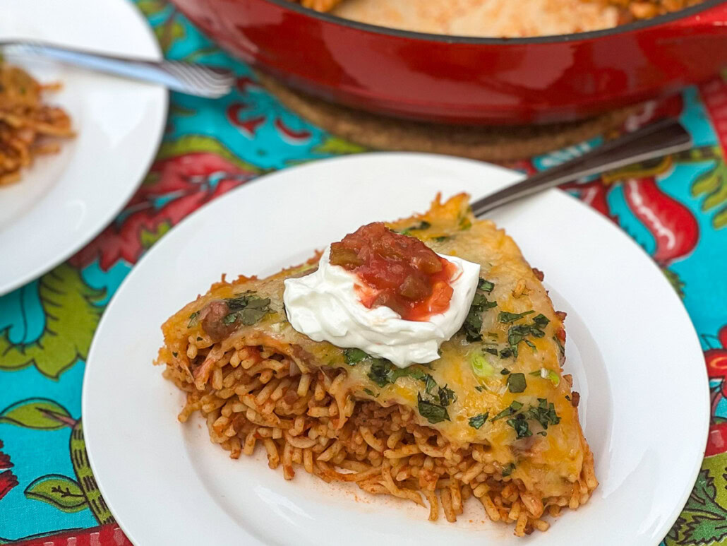 A slice of baked spaghetti with melted cheese, topped with sour cream and salsa, served on a white plate with a fork.
