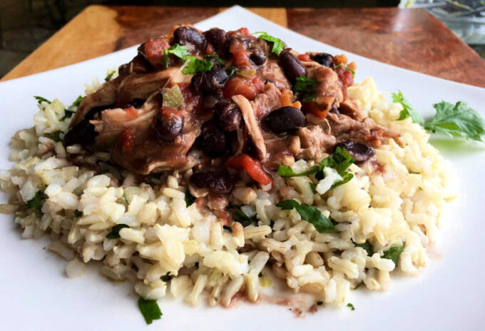 A serving of shredded chicken with black beans, tomatoes, and herbs over a bed of cooked rice, garnished with fresh parsley on a white plate.