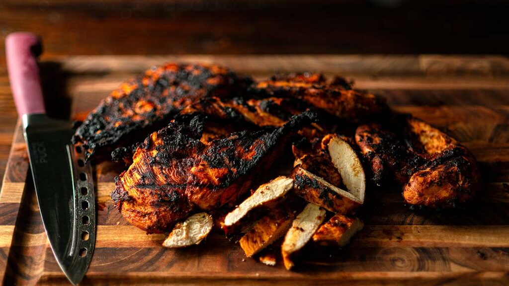 Grilled and slightly charred chicken breasts, some sliced, rest on a wooden cutting board next to a large kitchen knife with a maroon handle.