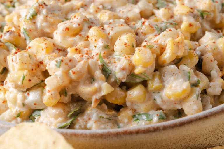 Close-up of a creamy corn salad with chopped herbs and a sprinkle of red seasoning, served in a ceramic bowl.