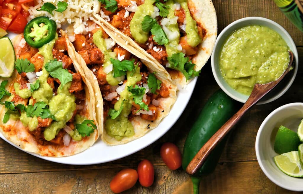 Three chicken tacos topped with guacamole, cilantro, cheese, and jalapeños on a plate, served with a bowl of green salsa, lime wedges, and grape tomatoes on a wooden table.