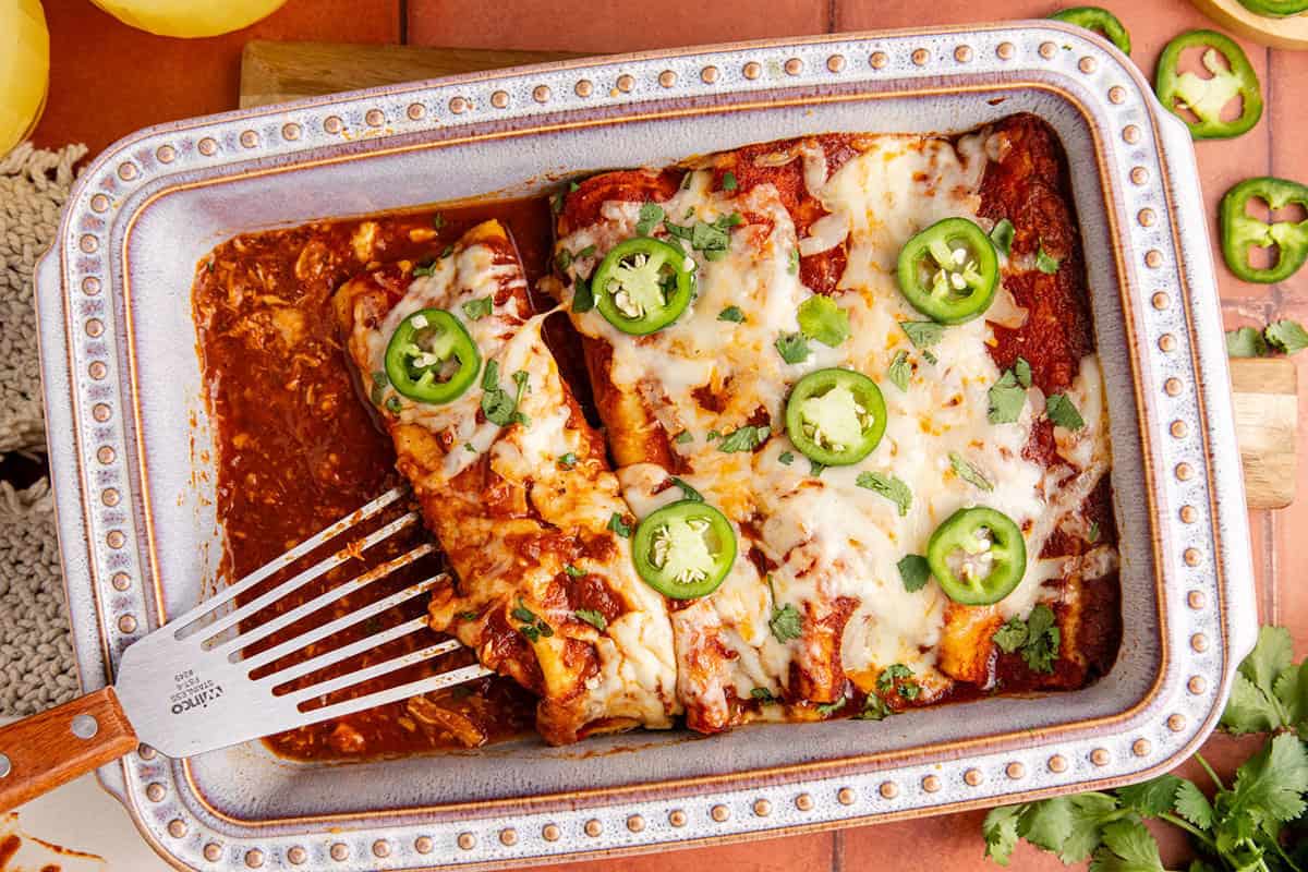 A baking dish with cheese-topped enchiladas garnished with sliced jalapeños and cilantro, with a serving spatula and one enchilada partially removed.