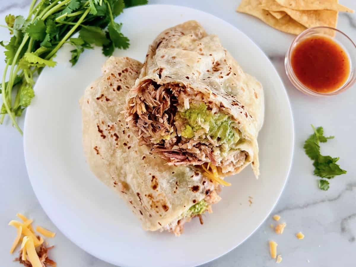 A burrito cut in half on a white plate, filled with shredded meat, rice, guacamole, and cheese, with cilantro, tortilla chips, and salsa on the side.