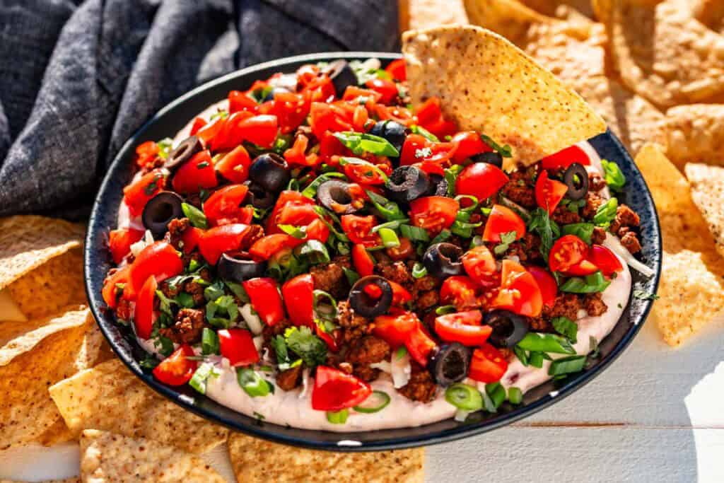 A plate of layered taco dip topped with chopped tomatoes, black olives, green onions, and cilantro, surrounded by tortilla chips.