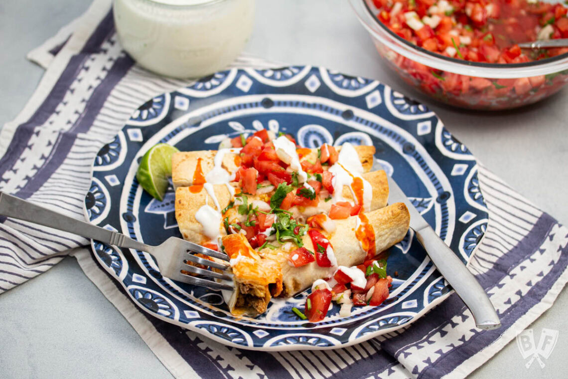 A plate with three enchiladas topped with pico de gallo and sour cream, served with a wedge of lime; fork and knife on the plate, with a bowl of salsa and a jar in the background.