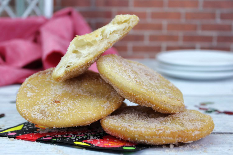 Four round sugared pastries are stacked on a colorful napkin; one pastry is broken in half, showing its fluffy interior.