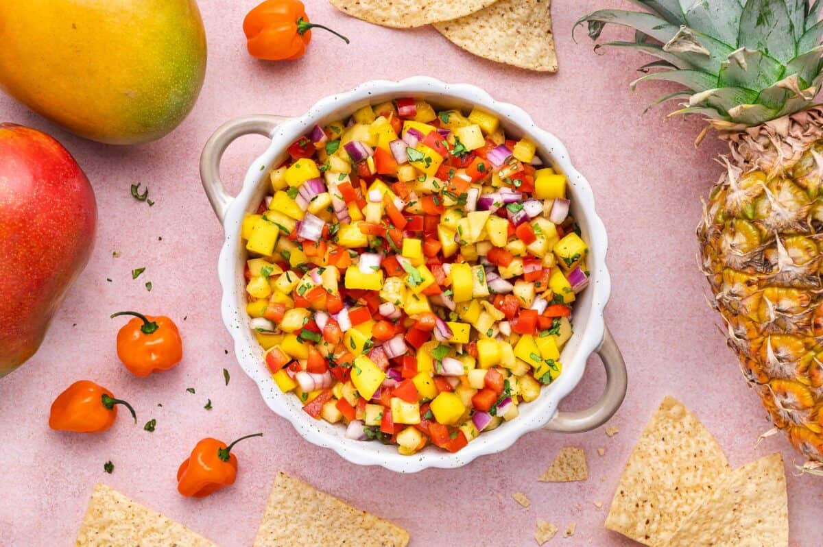 A bowl of mango salsa with diced mango, red onion, bell pepper, and herbs, surrounded by tortilla chips, mangoes, a pineapple, and habanero peppers on a pink surface.