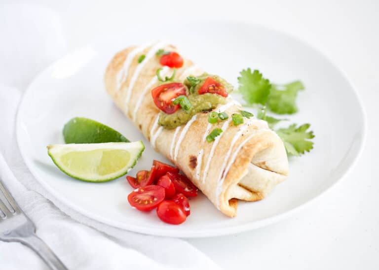 A baked burrito topped with sour cream, guacamole, diced tomatoes, and green onions, served with lime wedges, cherry tomatoes, and cilantro on a white plate.