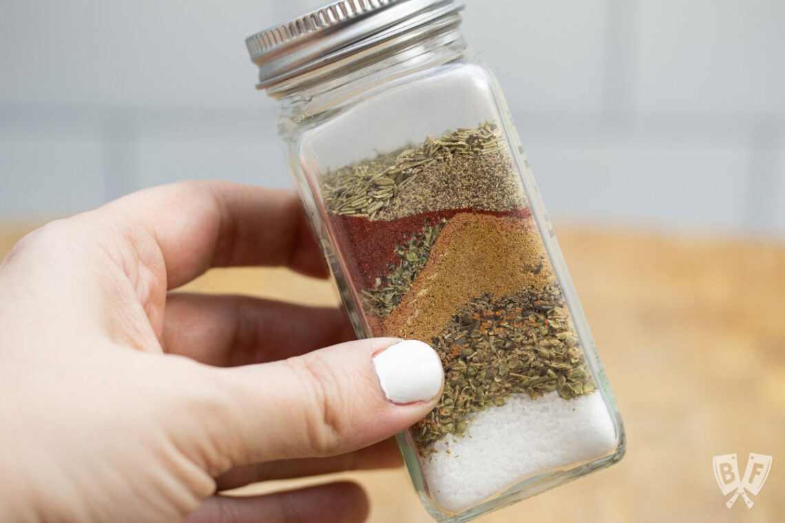 A hand holds a glass spice jar containing neatly layered spices and herbs, including salt, oregano, paprika, and pepper, with a metal lid.