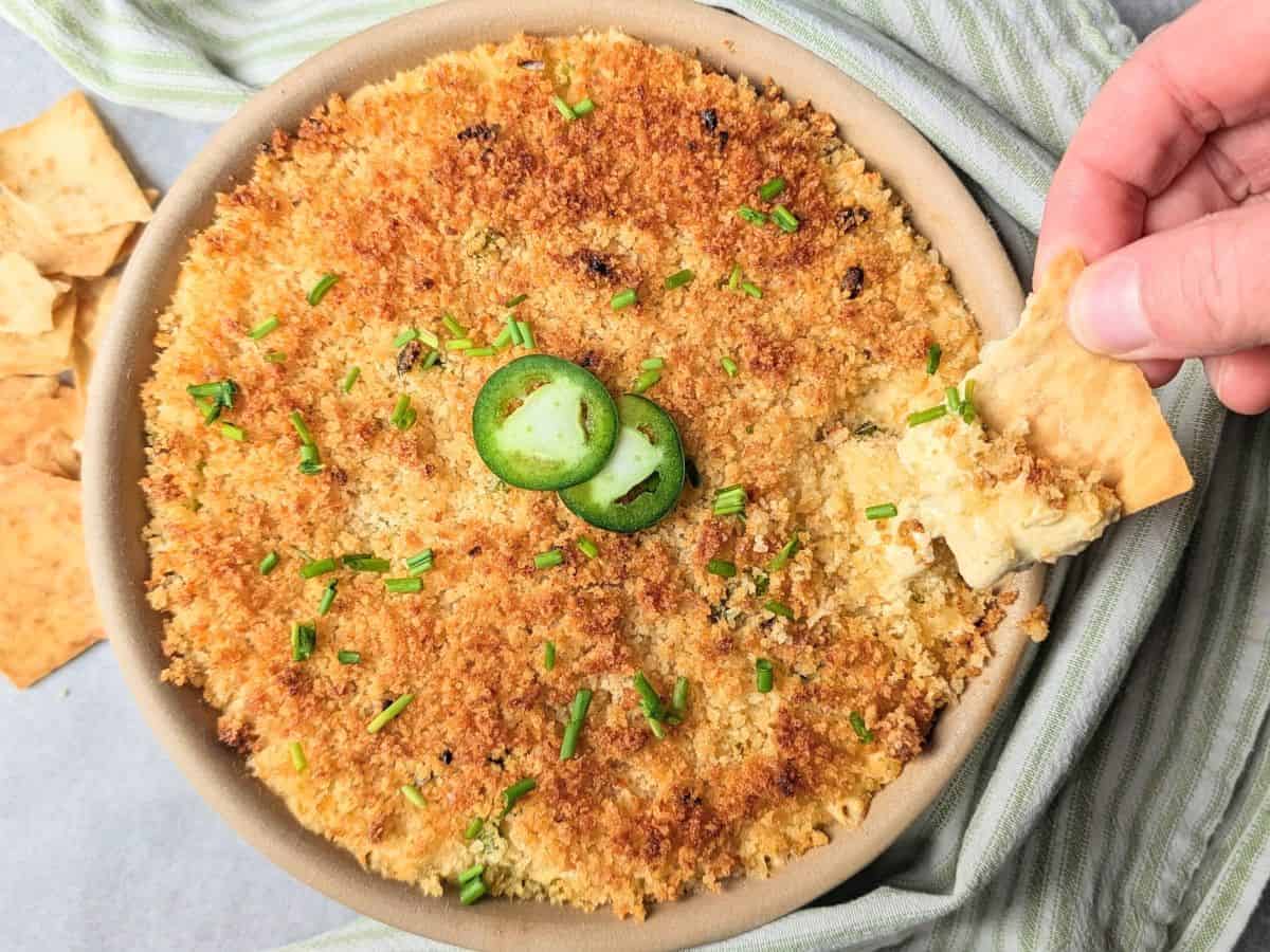 A hand dips a cracker into a baked dish topped with golden breadcrumbs, sliced jalapeños, and chopped chives.