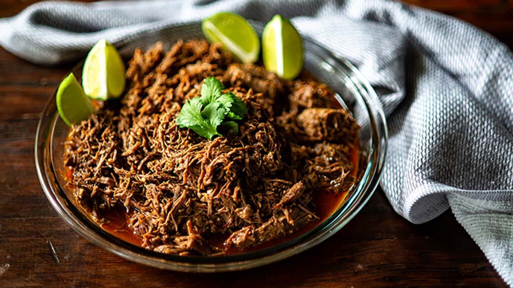 A glass dish filled with shredded beef in a red sauce, garnished with cilantro and lime wedges, with a gray cloth napkin beside it.