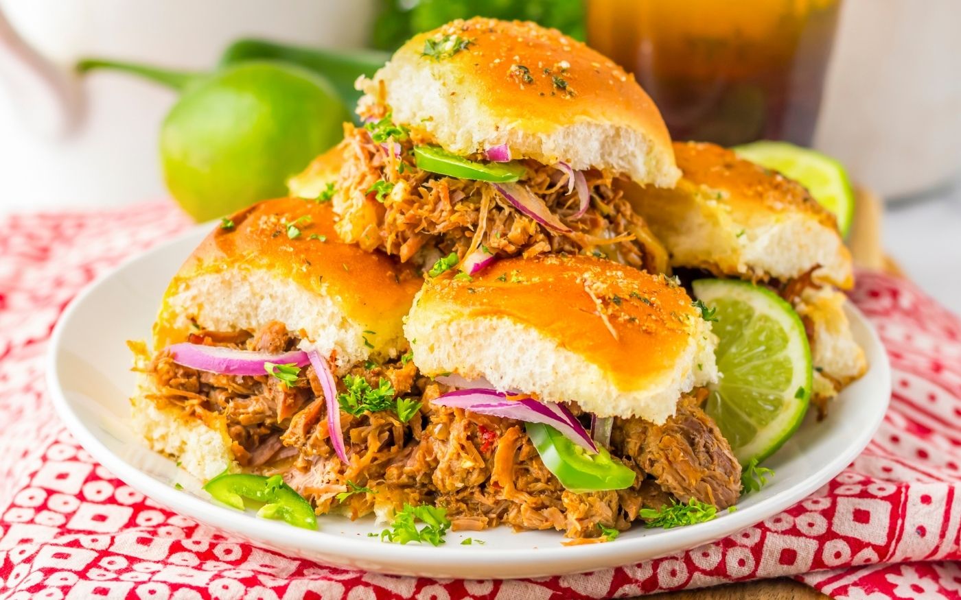 A plate of pulled pork sliders on soft buns, garnished with red onion, green peppers, herbs, and lime wedges, is set on a red and white patterned cloth.