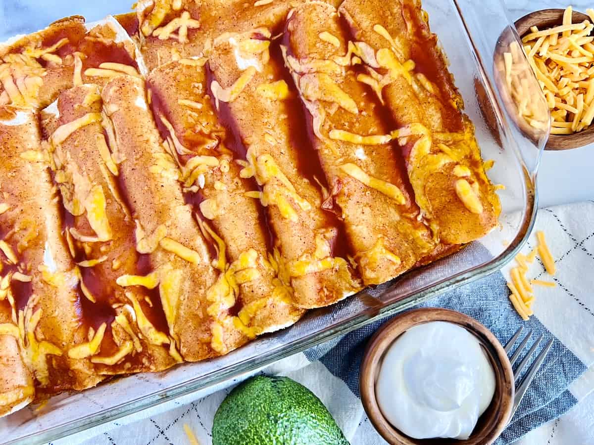 A glass baking dish with enchiladas topped with red sauce and shredded cheese, next to bowls of shredded cheese, sour cream, and a whole avocado.