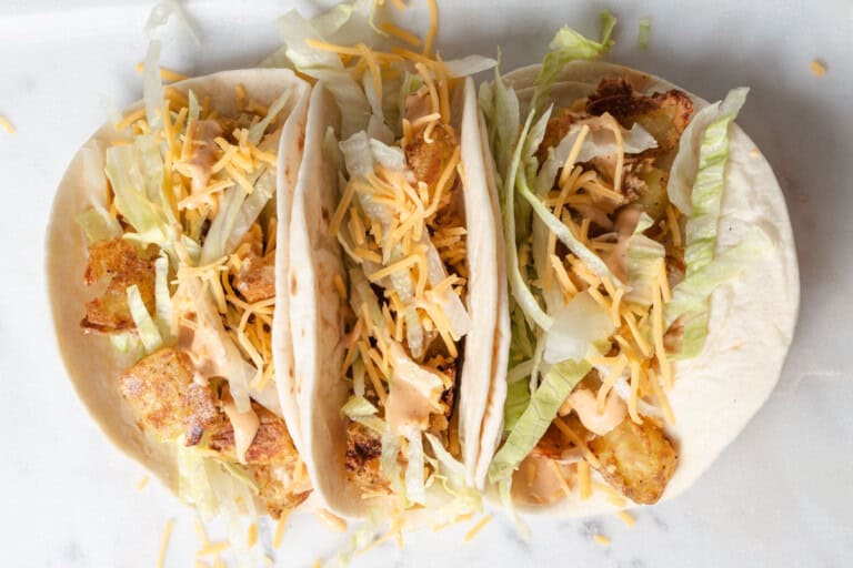 Three soft tacos filled with breaded fish, shredded lettuce, cheddar cheese, and a drizzle of creamy sauce on a white surface.