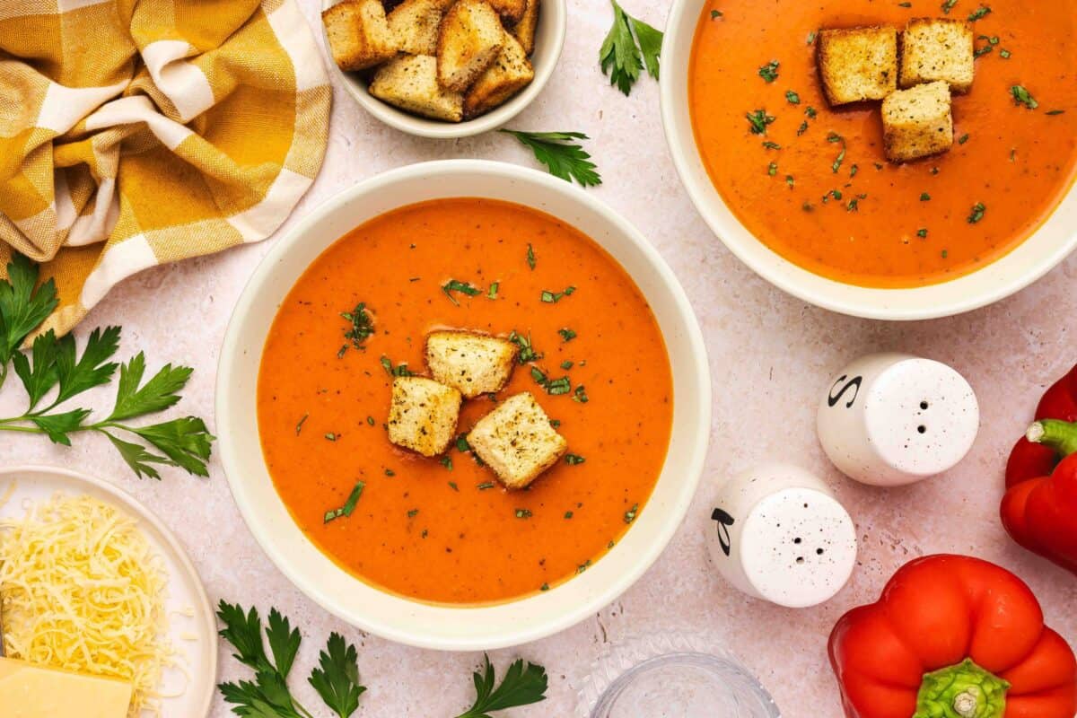 Two bowls of tomato soup topped with croutons, surrounded by parsley, shredded cheese, red bell pepper, salt and pepper shakers, and a yellow checkered napkin.