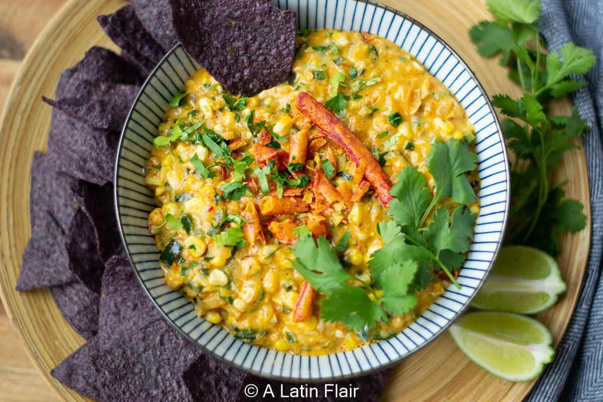 A bowl of creamy corn dip garnished with cilantro, cheese, and chili strips, surrounded by blue corn tortilla chips and lime wedges on a wooden platter.