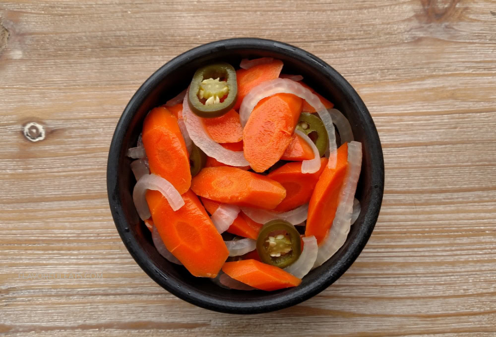 A black bowl filled with sliced carrots, onions, and jalapeños sits on a wooden surface.