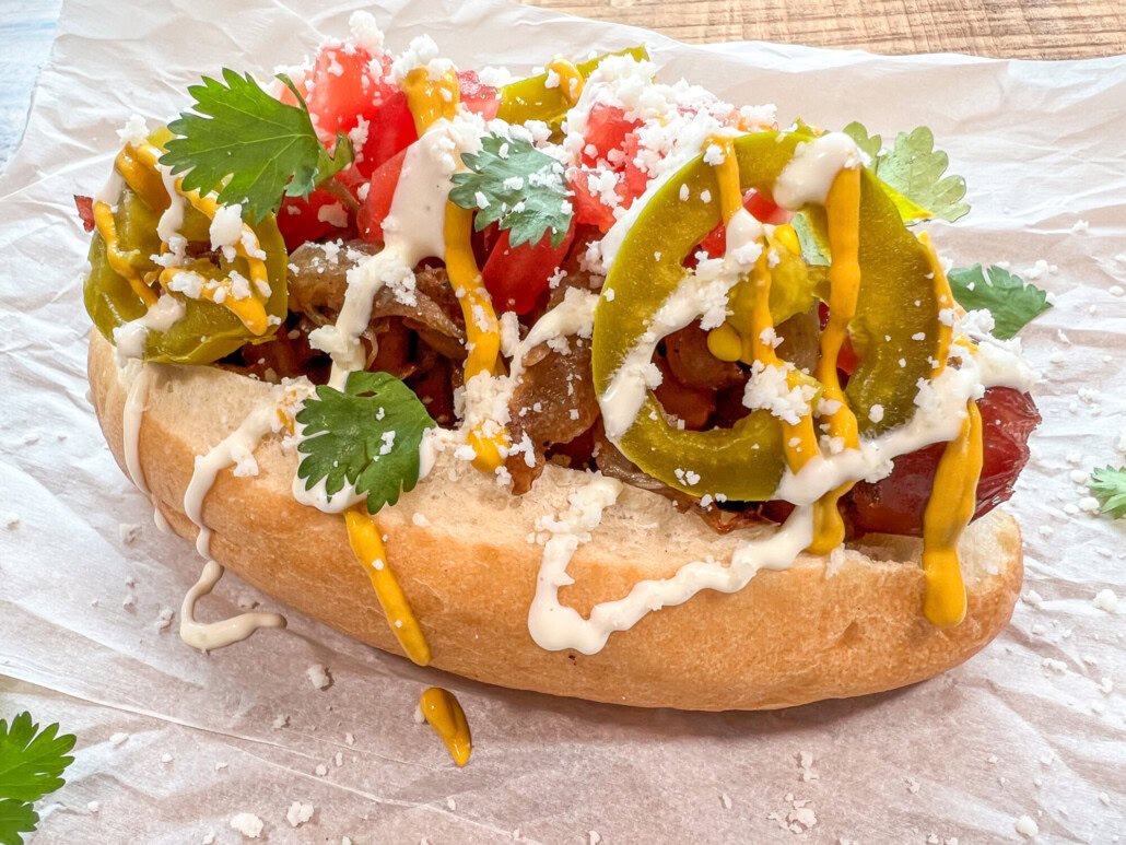 A hot dog topped with sliced jalapeños, chopped tomatoes, cilantro, crumbled cheese, mustard, and a white sauce, served on parchment paper.