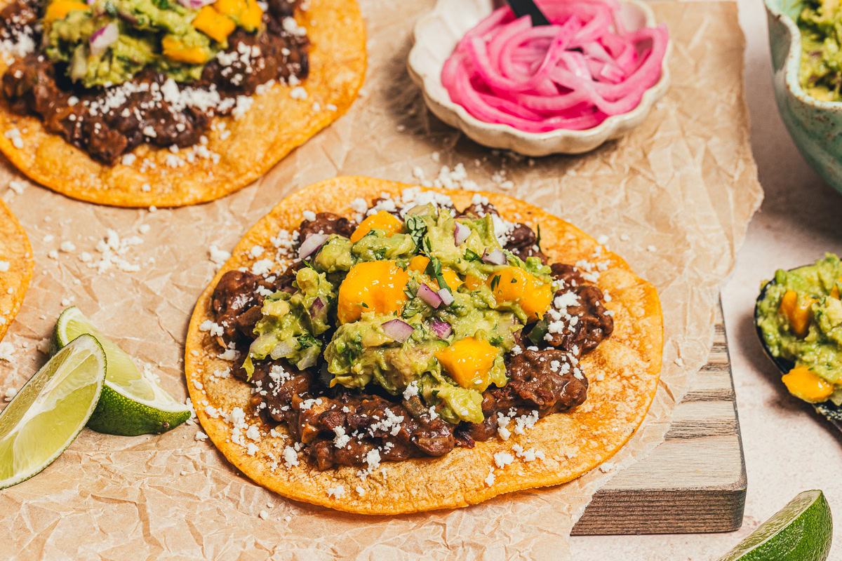 A corn tostada topped with black beans, crumbled cheese, guacamole with mango, and diced red onion, with lime wedges and pickled onions on the side.