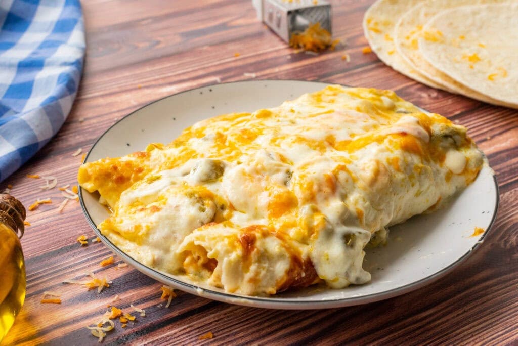 A plate of cheesy enchiladas covered in melted cheese sauce, with shredded cheese, tortillas, and a blue checkered napkin on a wooden table.