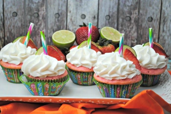 Five cupcakes with pink cake and white frosting are topped with lime and strawberry slices and colorful straws. Whole strawberries and limes are in the background.