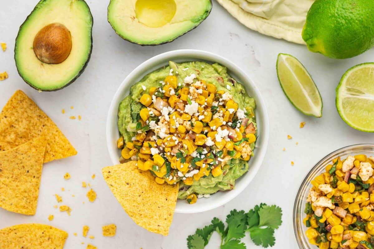 Bowl of guacamole topped with corn and crumbled cheese, surrounded by tortilla chips, avocado halves, lime wedges, cilantro, and a corn salad.