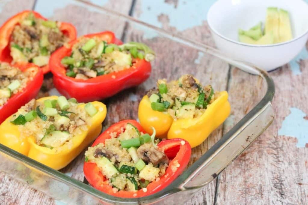 Red and yellow bell peppers halved and stuffed with a vegetable and grain mixture in a glass baking dish, with a small bowl of sliced avocado in the background.