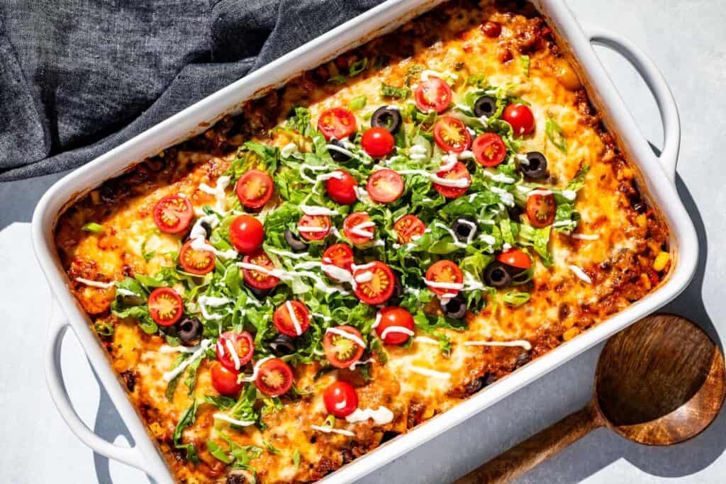 A baked casserole topped with melted cheese, lettuce, sliced cherry tomatoes, black olives, and a drizzle of white sauce in a white dish next to a wooden spoon.