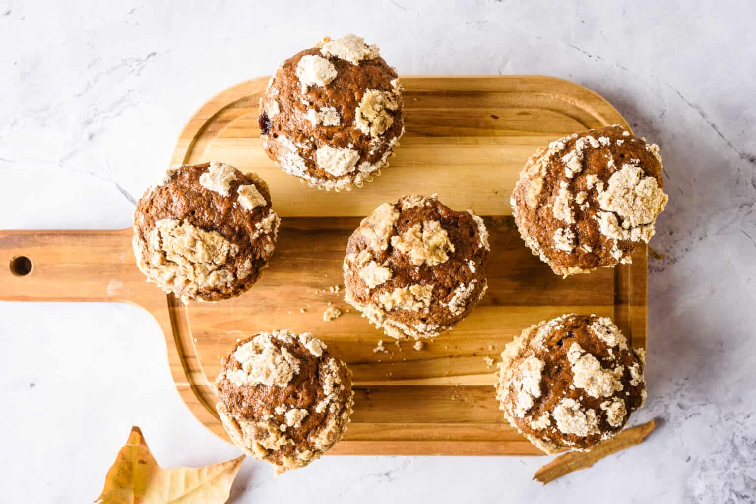 Five muffins with crumb topping arranged on a wooden cutting board, viewed from above, on a light marble surface with a leaf nearby.