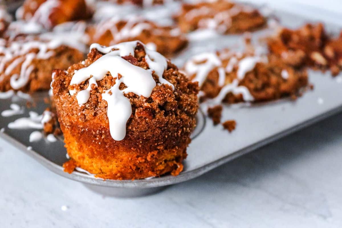 A muffin with crumb topping and white icing sits on the edge of a muffin tin, with more muffins in the background.