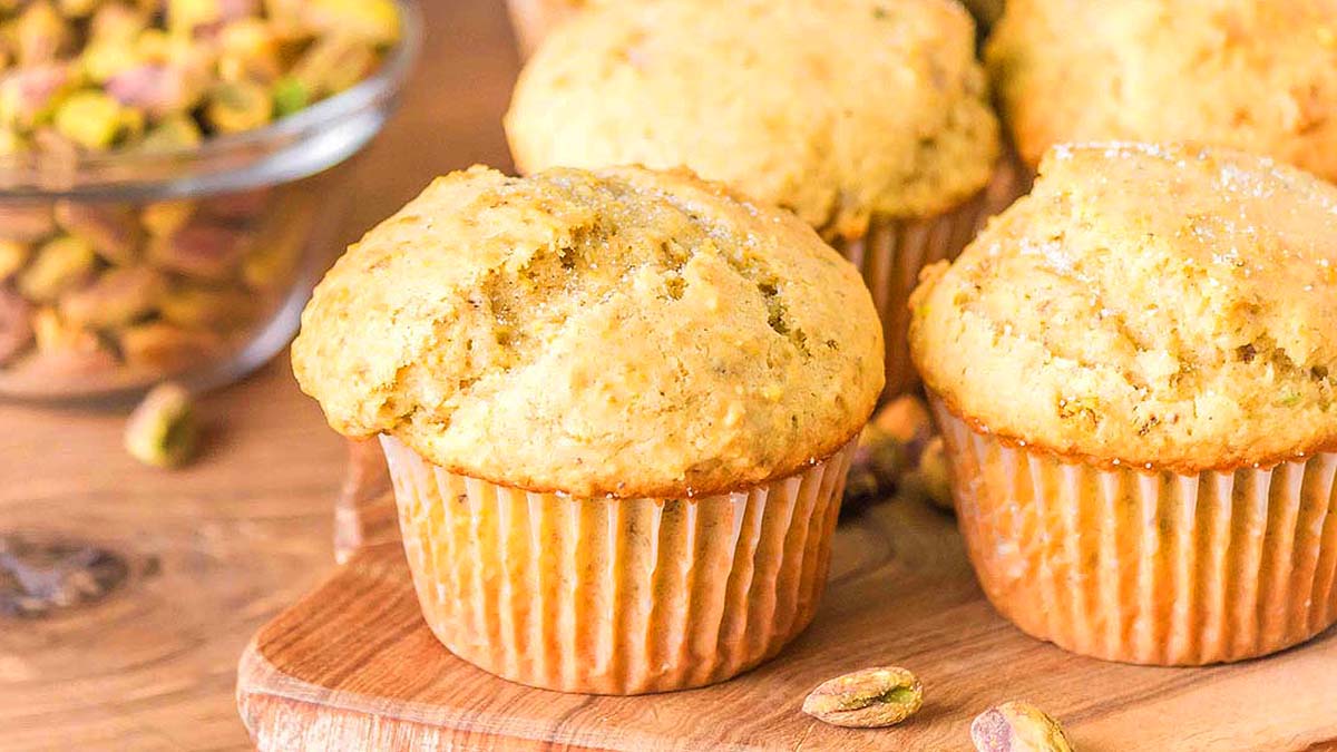 Three golden brown muffins on a wooden board with a bowl of pistachios and loose pistachios in the background.