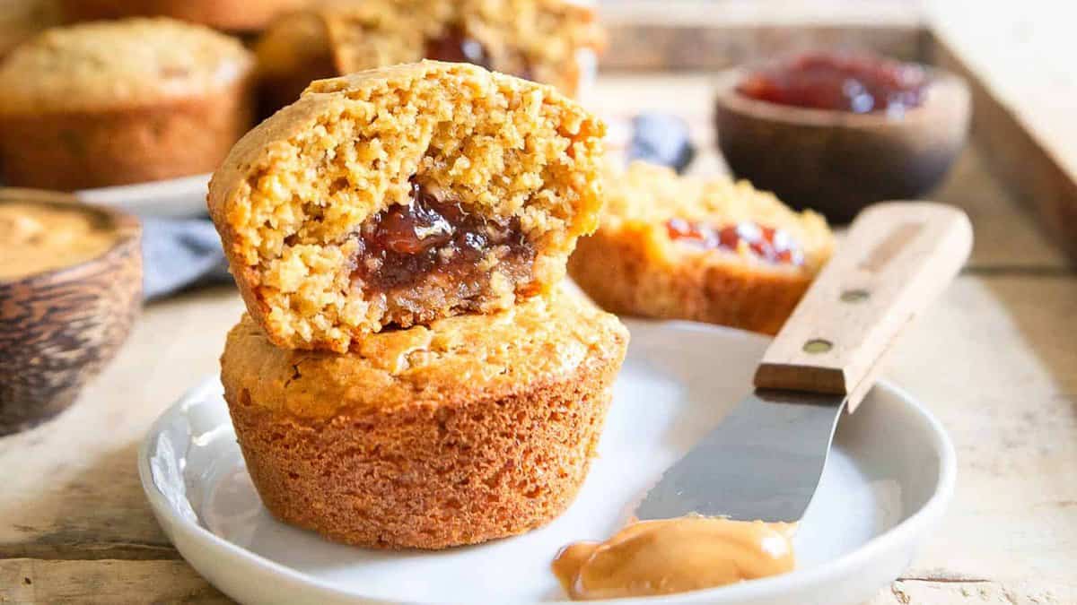 Two muffins are stacked on a white plate, with the top muffin cut open to show a peanut butter and jelly filling. A knife with peanut butter and a bowl of jelly are nearby.