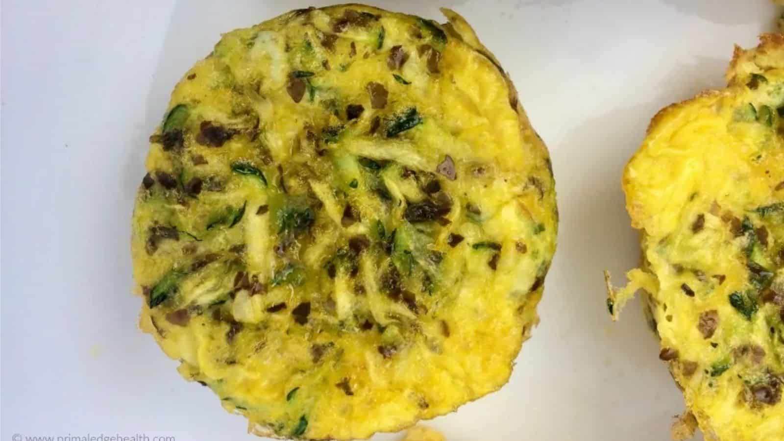 A round, baked egg and vegetable frittata sits on a white surface, showing visible pieces of green zucchini and herbs.