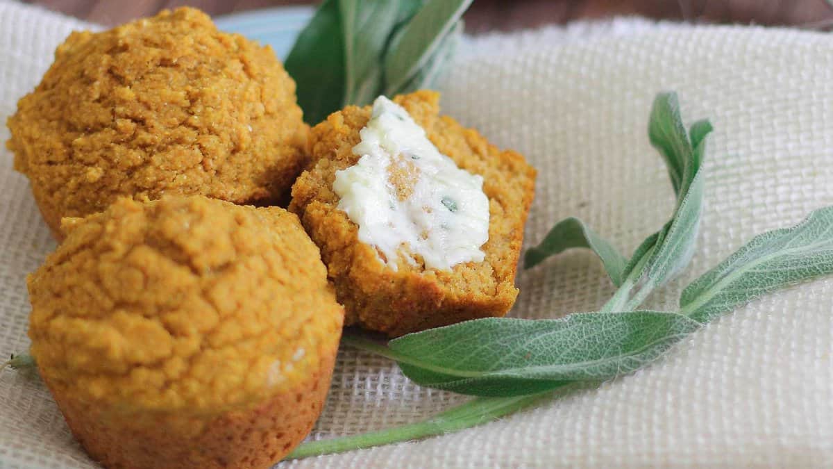 Three pumpkin muffins on a beige cloth, one split open showing a spread of butter, with fresh sage leaves beside them.