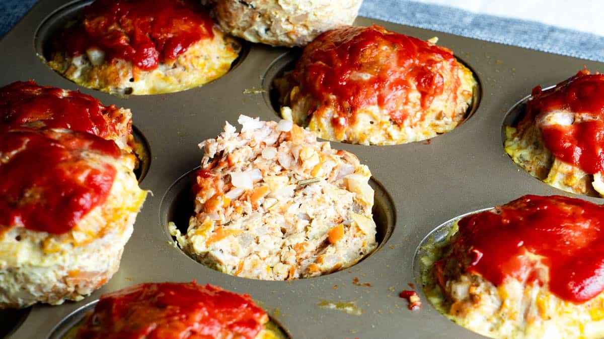 A muffin tin filled with individual meatloaf servings, each topped with ketchup; one meatloaf is cut open, showing its interior.