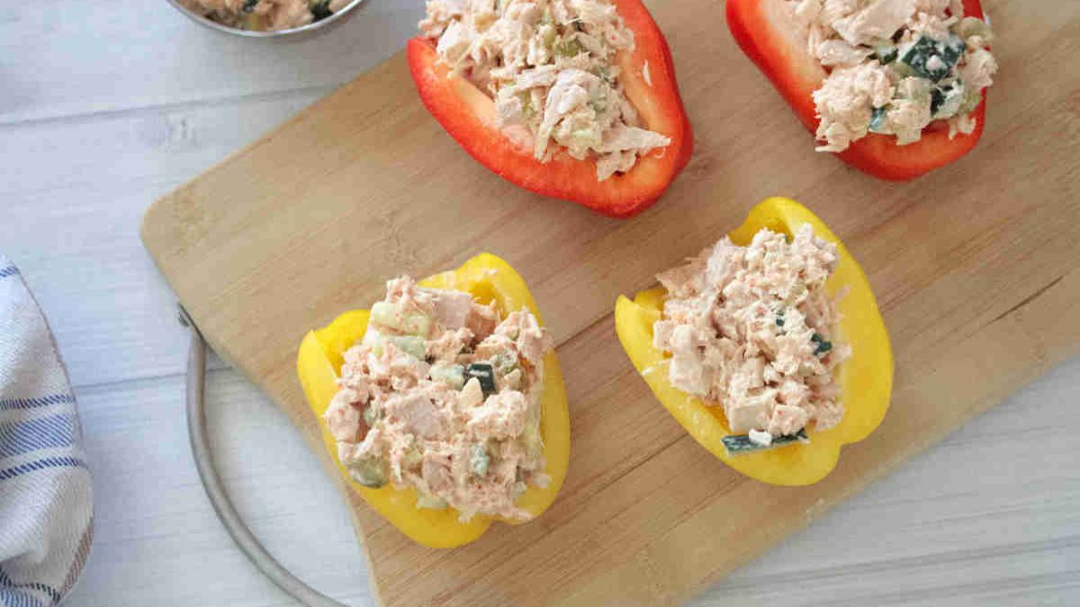 Halved red and yellow bell peppers filled with a chicken salad mixture sit on a wooden cutting board.