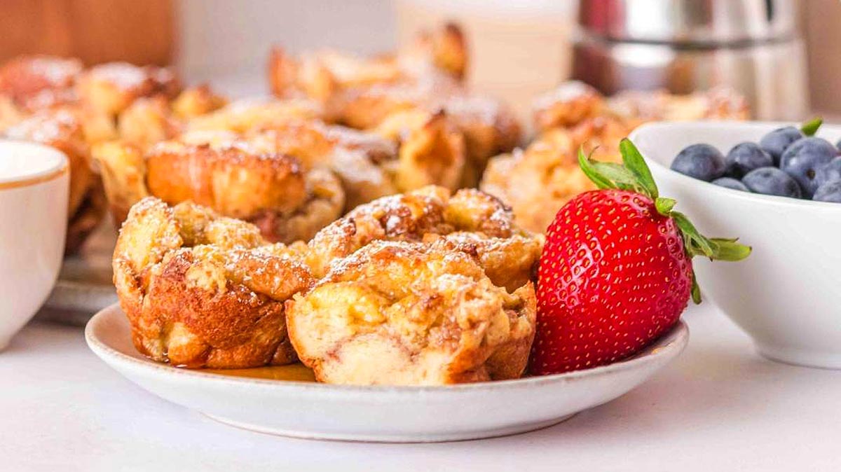 A plate with French toast muffins dusted with powdered sugar, served with a fresh strawberry and a bowl of blueberries on the side.