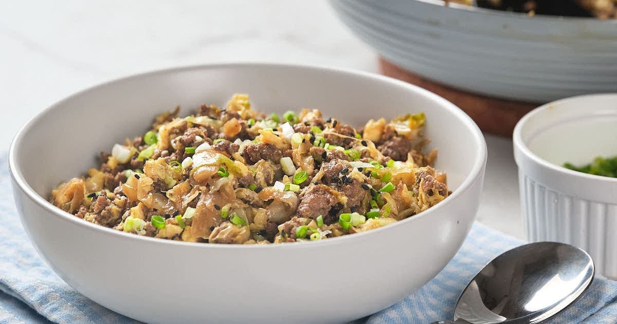 A white bowl filled with a mixture of ground meat, chopped cabbage, green onions, and sesame seeds sits on a blue cloth next to a spoon.
