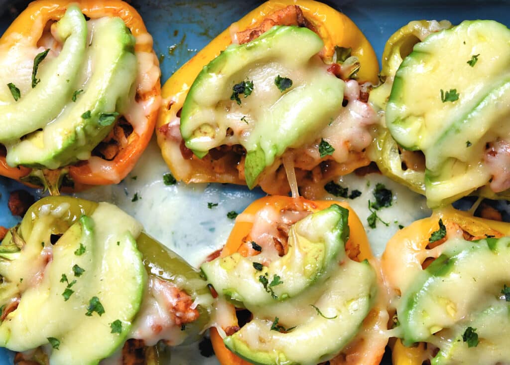 Yellow and green bell pepper halves stuffed with filling, topped with melted cheese and sliced avocado, garnished with chopped herbs.
