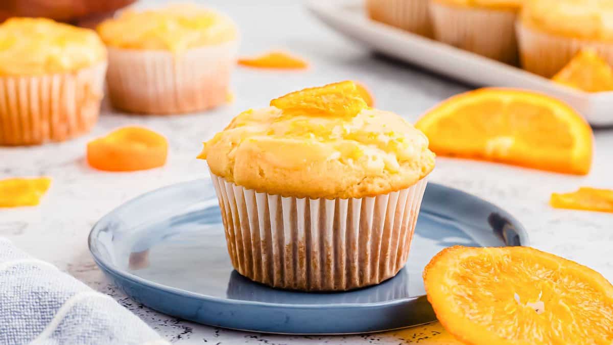 A single orange muffin on a blue plate, garnished with an orange slice, with more muffins and orange slices in the background.
