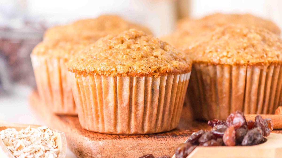Close-up of bran muffins on a wooden surface, with oats and raisins visible in the foreground.