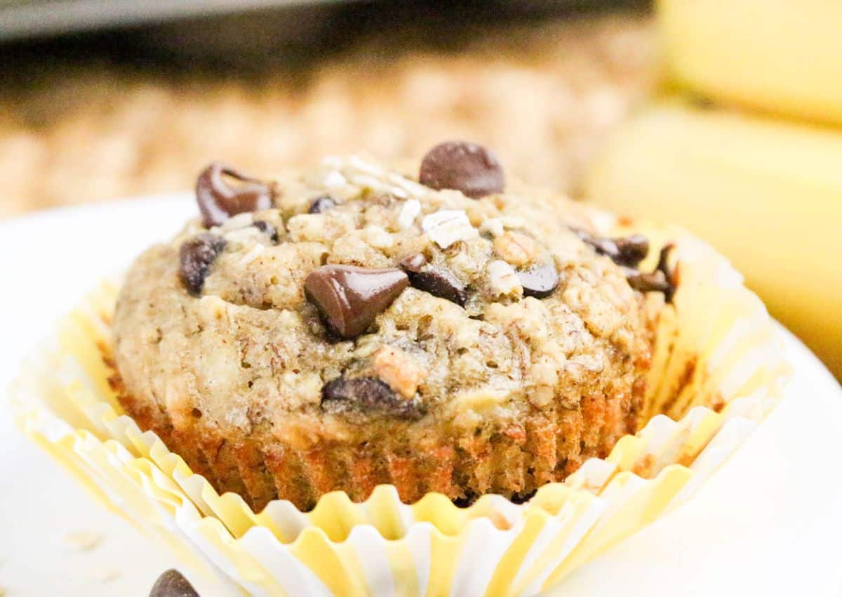 A close-up of a chocolate chip muffin in a yellow-striped paper liner, with chocolate chips and oats visible on top.