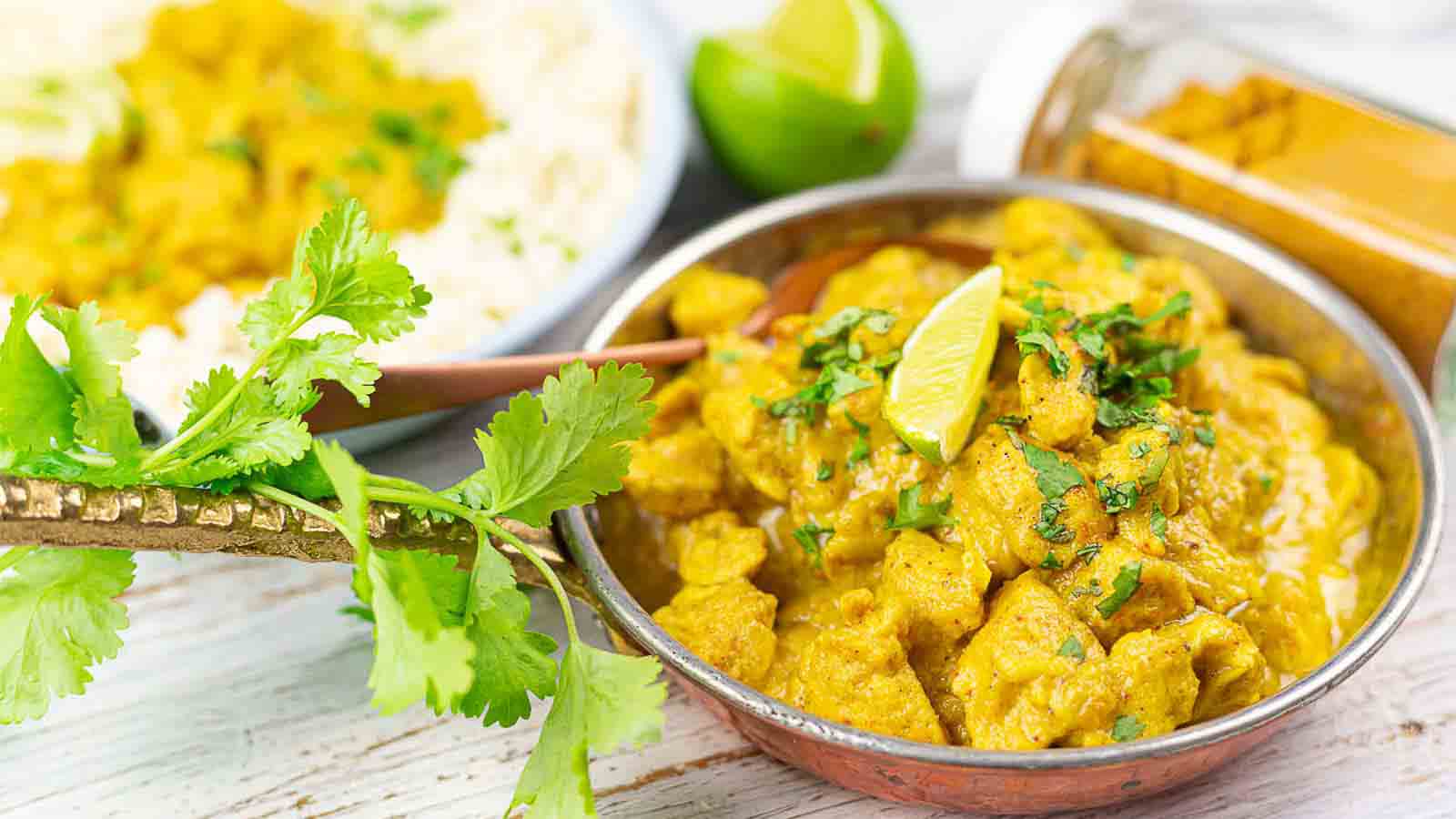A bowl of yellow curry with chicken, garnished with cilantro and a lime wedge, sits next to a plate of rice, fresh cilantro, and a jar of ground spices.