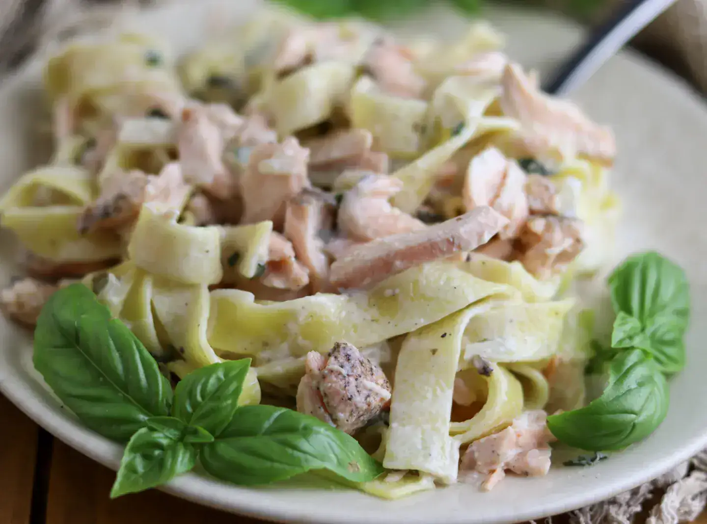A plate of fettuccine pasta with creamy sauce, chunks of salmon, and fresh basil leaves, with a fork on the side.