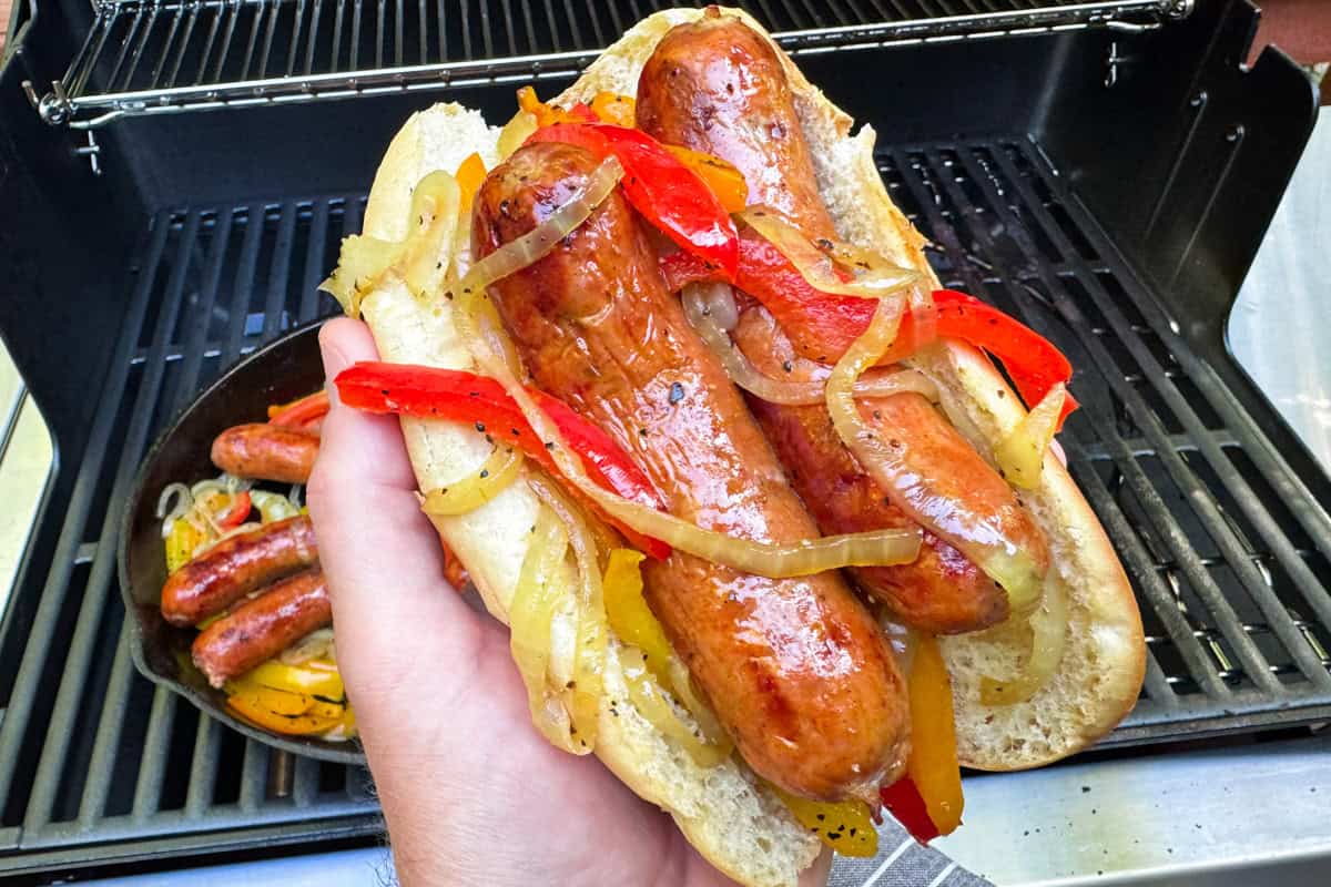 A hand holds a sausage sandwich with grilled onions and red peppers in front of a grill with more sausages cooking.
